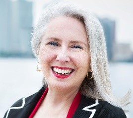 Woman with shoulder length blonde hair wearing a suit jacket and smiling with buildings and the Hudson River in the background.