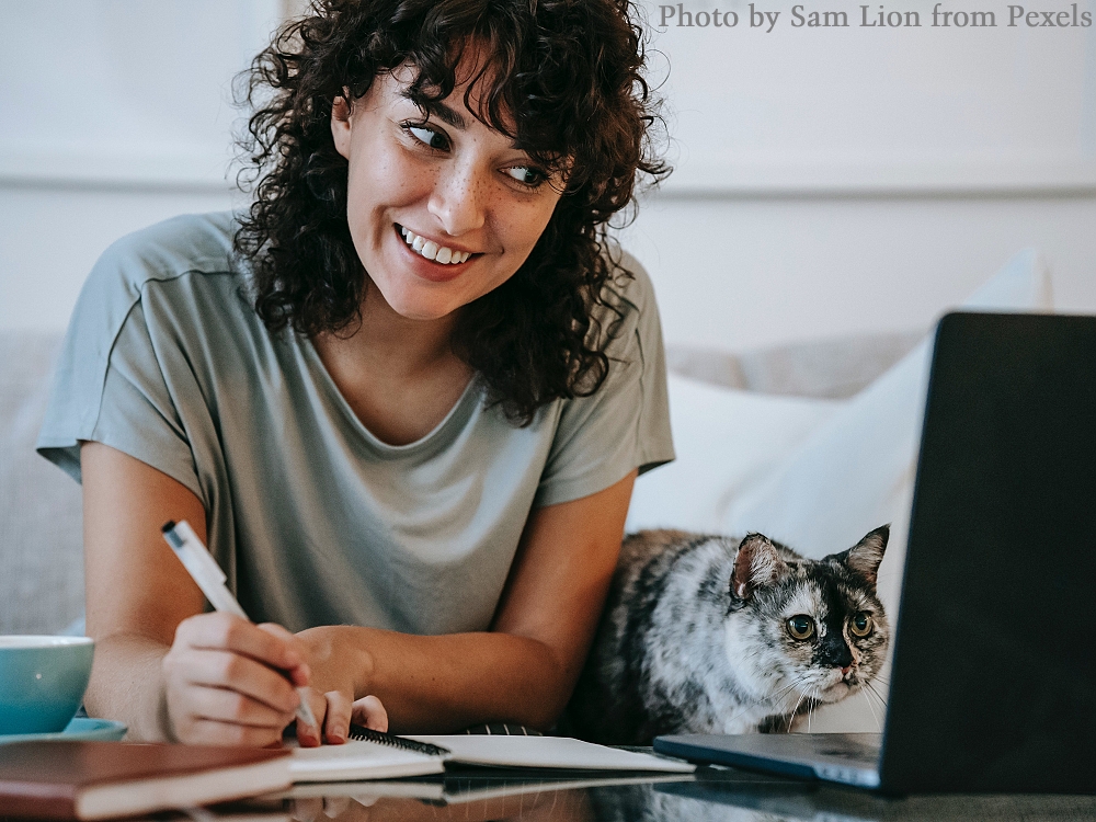 A woman writing in a notebook while her cat looks at the computer screen.